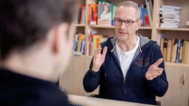 Das Bild zeigt Clemens Hillenbrand im Gespräch mit dem Interviewer. Hillenbrand sitzt vor einem Bücherregal und gestikuliert.